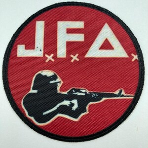 Puede incluir: Un parche bordado circular con fondo rojo y borde negro. El parche presenta el texto blanco J.F.A. sobre una silueta negra de una persona con casco apuntando con un rifle. Pequeñas cruces blancas están situadas entre las letras.