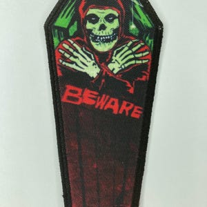 May include: A coffin-shaped embroidered patch featuring a green skull wearing a red hooded cloak with arms crossed. The word BEWARE is printed in red capital letters below the skull. The background includes green and black tones with a spiderweb.
