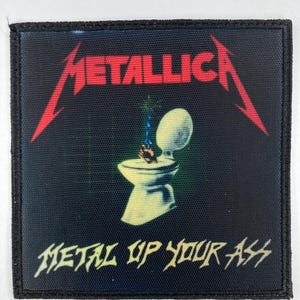Peut inclure: Un écusson noir avec le nom du groupe "Metallica" en rouge, lettrage stylisé. En dessous, une image de toilettes avec une flamme. Le texte "Metal Up Your Ass" est en dessous.