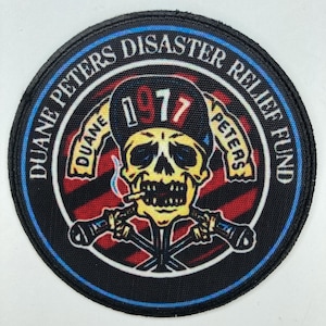 Puede incluir: Un parche bordado circular que presenta una calavera amarilla con una gorra negra con el texto 1977. El diseño incluye dos micrófonos cruzados, un cigarrillo y el texto DUANE PETERS DISASTER RELIEF FUND en un borde negro con ribete azul.