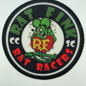 Può includere: Una toppa circolare ricamata raffigurante un topo dei cartoni animati verde con una maglietta rossa e le lettere RF. Il bordo nero contiene il testo verde RAT FINK e il testo rosso RAT RACERS, con le lettere bianche CC e SC sui lati.