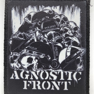 May include: A rectangular black and white fabric patch featuring a graphic illustration of multiple skulls wearing police helmets with the words POLIZEI and POLICJA visible. The text AGNOSTIC FRONT is printed in large white letters at the bottom.