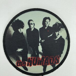 May include: A round black and white patch with a band of black stitching around the edge. The patch features a black and white photo of a band with the text "The Humans" in red letters.