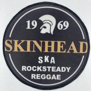 May include: A round black patch with white and gold text. The patch features the words "SKINHEAD" and "SKA ROCKSTEADY REGGAE". The year "1969" is also displayed, along with a white helmet graphic.
