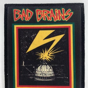 Pode incluir: Patch quadrado preto com as palavras "BAD BRAINS" em vermelho, contornadas em amarelo. O patch apresenta um raio amarelo sobre uma cúpula branca com fundo preto, emoldurado por listras vermelhas, amarelas e verdes.