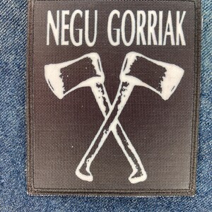 Puede incluir: Un parche rectangular de tela negra con el texto blanco NEGU GORRIAK en la parte superior y una ilustración gráfica de dos hachas cruzadas en blanco debajo. El parche tiene un borde cosido y se muestra sobre un fondo de tela vaquera azul.