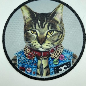 Puede incluir: Un parche bordado circular que muestra un gato atigrado marrón con un chaleco de mezclilla azul con tachuelas plateadas, un cuello con estampado de leopardo y un collar rojo con un dije de pluma. El chaleco tiene botones con The Clash y Catnip.
