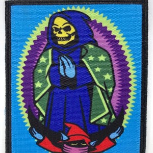 Op de afbeelding: Vierkante patch met een cartoon illustratie van een skeletfiguur in een blauwe jurk en groene cape met steraccenten, tegen een blauwe achtergrond. Het ontwerp bevat een rode banner met een personage en een rode hoed.