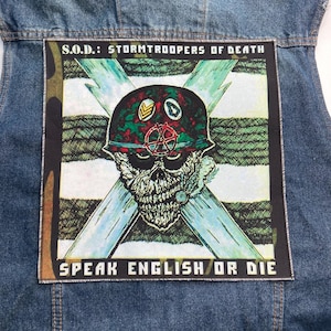 May include: A blue denim vest featuring a large square patch on the back. The patch displays a graphic of a skull wearing a camouflage helmet with a red symbol, set against a white and green striped background with the text S.O.D.: STORMTROOPERS OF DEATH and SPEAK ENGLISH OR DIE.