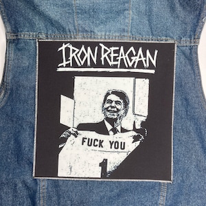 Puede incluir: Un chaleco vaquero azul con un parche negro grande. El parche muestra el nombre de la banda "IRON REAGAN" en blanco, encima de una imagen en blanco y negro de una persona sosteniendo una camiseta con las palabras "FUCK YOU" y el número "1".