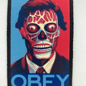 Könnte beinhalten: Ein rechteckiger Aufnäher mit einem stilisierten Bild eines schädelartigen Gesichts mit roten und weißen Details vor einem blauen und roten Hintergrund. Das Wort "OBEY" ist unten in Blau aufgedruckt. Der Aufnäher hat einen schwarzen Rand.