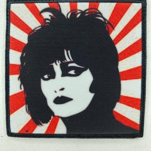 May include: A square patch with a black and white graphic of a woman's face. The background is a red and white striped pattern. The patch has a black border.