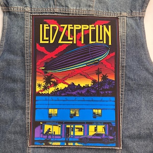 May include: A blue denim jacket featuring a large rectangular patch on the back. The patch displays a Led Zeppelin graphic with a purple airship, red clouds, a sunset, palm trees, and a blue building labeled Kingswell Los Feliz with a car.