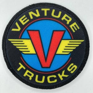 Könnte beinhalten: Runder Aufnäher mit dem gelben Schriftzug "VENTURE TRUCKS", der sich um einen blauen Kreis wölbt. Ein rotes "V" befindet sich in der Mitte, mit gelben Flügeln auf beiden Seiten. Der Aufnäher hat einen schwarzen Rand.