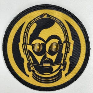 Puede incluir: Un parche redondo con un diseño negro y amarillo de la cara de un robot. La cara del robot es negra, con detalles amarillos y un borde negro. Adecuado para ropa o accesorios.