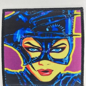 Puede incluir: Parche cuadrado con una ilustración estilizada de un personaje con máscara y disfraz negros. La imagen tiene una cara amarilla, labios rojos y ojos azules, sobre un fondo morado y negro.