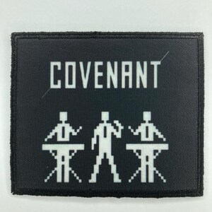 May include: A rectangular black patch with a white embroidered design. The text COVENANT is printed in a bold, blocky font above three pixelated figures. Two figures stand behind consoles, while the central figure stands between them.