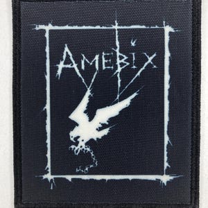 Puede incluir: Un parche cuadrado negro con la palabra "AMEBIX" en blanco, con una fuente estilizada, y un gráfico blanco de un pájaro o criatura alada. El diseño está enmarcado por un borde blanco y un borde negro.