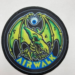 Puede incluir: Un parche redondo con borde negro que presenta una criatura alada verde y amarilla con un solo ojo azul. La palabra "AIRWALK" se muestra en letras azules sobre un fondo azul.