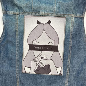 Puede incluir: Un chaleco vaquero azul claro con un parche rectangular en la espalda. El parche presenta una ilustración en blanco y negro de un personaje de dibujos animados con la palabra "Wonderland" sobre los ojos.