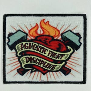 Puede incluir: Un parche rectangular con borde negro presenta un corazón atravesado por dos hachas, con llamas encima. Una pancarta sobre el corazón dice "AGNOSTIC FRONT DISCIPLINE" en texto negro. El fondo es blanco con rayos rojos.