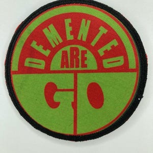 Può includere: Una toppa ricamata circolare con un bordo nero cucito. Il design presenta un semicerchio superiore rosso con il testo DEMENTED in verde, una barra orizzontale centrale rossa con il testo ARE in verde e una sezione inferiore verde con il testo GO in rosso.