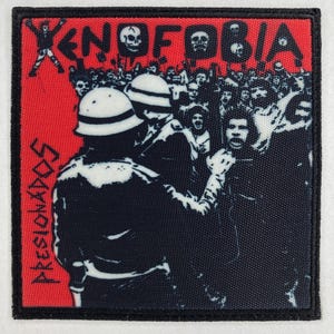 Puede incluir: Parche cuadrado con fondo rojo y borde negro. El parche presenta la palabra "XENOFOBIA" sobre una ilustración de una multitud. La palabra "PRESIONADOS" está impresa verticalmente en el lado izquierdo. La imagen representa una escena de protesta.