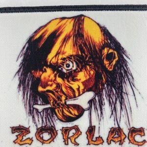 Puede incluir: Un parche blanco con borde negro que presenta un gráfico de una cara grotesca con tonos naranja y amarillo, sosteniendo un hueso. El texto "ZORLAC SKATEBOARDS" está impreso en naranja.