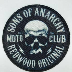 Puede incluir: Un parche bordado negro de forma circular que presenta una calavera blanca con gafas de sol en el centro. El texto dice SONS OF ANARCHY MOTO CLUB REDWOOD ORIGINAL, con EST y 67 a ambos lados de la calavera y dos pequeños rayos.