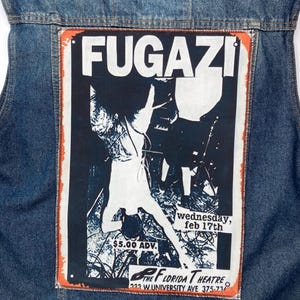May include: A blue denim jacket featuring a large rectangular patch on the back. The patch displays a black and white concert poster for Fugazi at The Florida Theatre, including the text "wednesday, feb 17th", "$5.00 ADV.", and the venue address.
