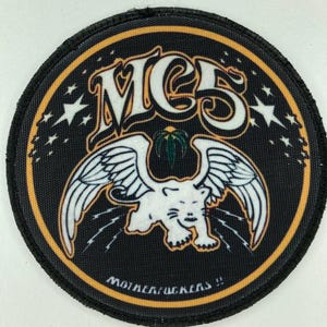 Puede incluir: Parche redondo negro con un diseño de tigre alado blanco y el texto "MCS" en una fuente estilizada. El parche tiene un borde amarillo y el texto "MOTHERFUCKERS !!" en la parte inferior.