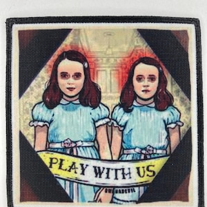 Könnte beinhalten: Ein quadratischer Aufnäher mit zwei Mädchen in hellblauen Kleidern, die sich an den Händen halten. Die Mädchen haben dunkles Haar und sind in einer Raute zentriert. Die Worte "PLAY WITH US" stehen auf einem gelben Banner unten.