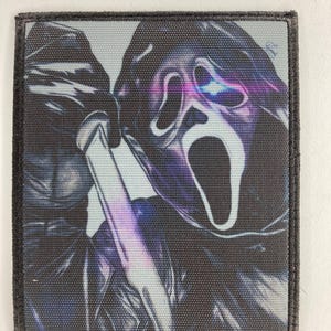 Puede incluir: Parche cuadrado con una imagen estilizada de la máscara de Ghostface de la serie de películas de terror Scream. El diseño incluye un cuchillo y un fondo oscuro texturizado con reflejos morados y azules. El parche tiene un borde negro.