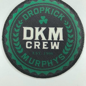 May include: A circular embroidered patch with a black border and a green serrated edge. The centre features the white text DKM CREW, a small green shamrock, and the text MURPHYS EST. 1996 in a circular pattern with green laurel leaf accents.