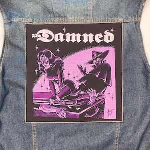 Puede incluir: Un chaleco vaquero azul con un parche rectangular. El parche muestra el nombre de la banda "The Damned" en letras góticas blancas. La ilustración es en morado y negro, con dos figuras al estilo cómic.