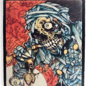 Puede incluir: Parche cuadrado con una ilustración detallada de una figura similar a una momia con un compañero esquelético. La obra de arte presenta una paleta de colores de azules, rojos y amarillos, con un borde negro.