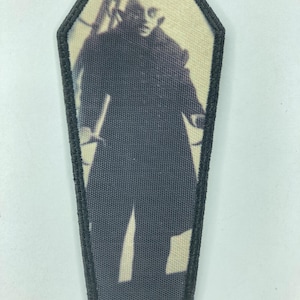 Op de afbeelding: Een doodskistvormige patch met een zwarte rand. De patch heeft een grijswaardenafbeelding van een figuur in een lange jas. De afbeelding heeft een vintage, enigszins wazig uiterlijk, wat wijst op een oudere foto of illustratie. Het algemene thema is gotisch of horror.