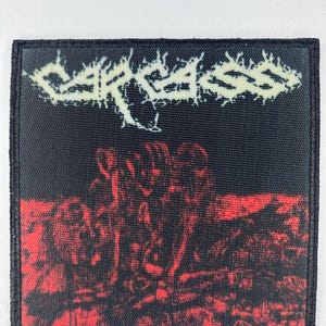 May include: Black and red patch featuring the band name "CARCASS" in a distressed, off-white font. The bottom half of the patch displays a red and black graphic. The patch has a black border.