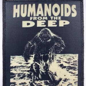 Puede incluir: Un parche bordado en blanco y negro con el texto "Humanoids From The Deep" y un gráfico de una criatura de las profundidades emergiendo del agua.