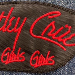 May include: Black patch with red embroidered text that reads "Hey Cric" above "Girls Girls" on a denim background. The patch has a black border and is likely a decorative element for clothing.