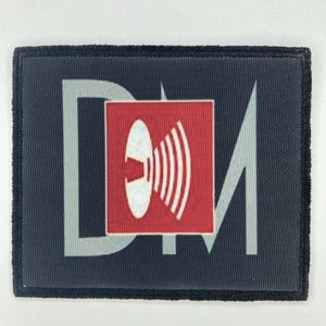 Puede incluir: Un parche rectangular de tela con fondo gris oscuro que presenta las letras grandes de color gris claro DM. Centrado sobre las letras hay un cuadrado rojo que contiene un gráfico blanco de un disco circular que emite ondas sonoras curvas hacia la derecha.