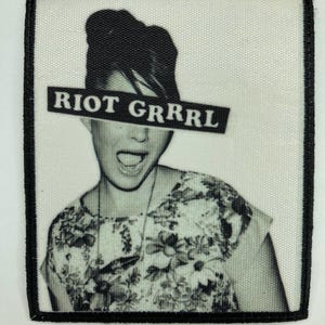 Könnte beinhalten: Ein quadratischer Stoffaufnäher mit einem schwarz-weißen Fotodruck einer Person mit offenem Mund. Ein schwarzer rechteckiger Balken mit dem weißen Text "RIOT GRRRL" verdeckt die Augen. Der Aufnäher hat einen dicken, schwarz umstickten Rand.
