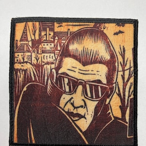 May include: A brown and black embroidered patch featuring a cartoon illustration of a person wearing sunglasses and a dark jacket. The person is looking to the side and has a serious expression. The background of the patch is a light brown color and features a cartoon illustration of a town.