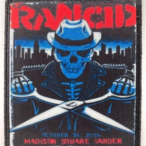 Może przedstawiać: Kwadratowa naszywka z niebieską czaszką w fedorze, trzymającą nożyczki, z nazwą zespołu "RANCID" w kolorze czerwonym. Widoczny jest również tekst "OCTOBER 19, 2019 MADISON SQUARE GARDEN NEW YORK CITY". Naszywka ma czarną obwódkę.