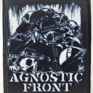 Puede incluir: Parche en blanco y negro con un diseño de calaveras y cascos, con el texto "AGNOSTIC FRONT". Los cascos llevan las palabras "POLIZEI" y "POLICJA". El parche tiene un borde negro.