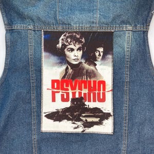Pode incluir: Um colete de ganga azul com um remendo vintage do cartaz do filme "Psicose" costurado nas costas. O remendo apresenta uma imagem em preto e branco de uma mulher e um homem com o título "Psicose" em letras vermelhas.