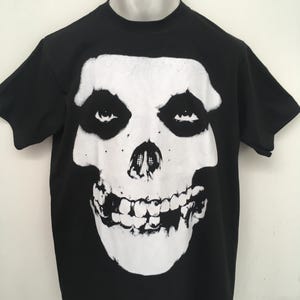 Puede incluir: Camiseta negra con un gráfico de calavera blanca. La calavera tiene una gran sonrisa con dientes y ojos negros.