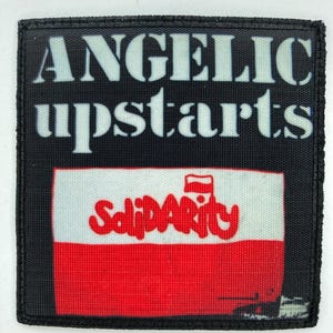 Puede incluir: Un parche negro con las palabras "ANGELIC upstarts" en blanco. Debajo, un rectángulo rojo y blanco muestra la palabra "Solidarity" en escritura roja. El parche tiene un borde negro.