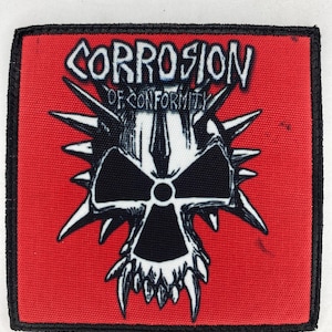 Op de afbeelding: Een rechthoekige patch met een rode achtergrond en een zwarte rand. De patch heeft een zwart-wit schedelontwerp met spikes en de tekst "CORROSION OF CONFORMITY" in witte, gestileerde letters.