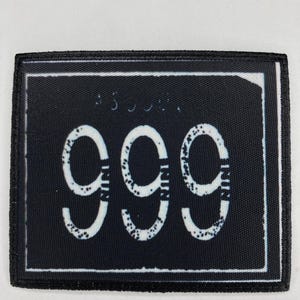Puede incluir: Un parche rectangular de tela negra con el número 999 en grandes dígitos blancos. Los dígitos tienen una textura desgastada y moteada. El parche está bordeado por una fina línea blanca y un borde cosido negro grueso y definido.